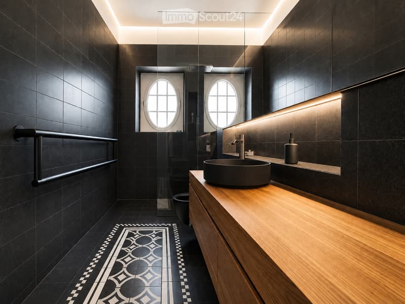 Modern bathroom with dark tiles, wooden vanity, round sink, sleek faucet, and built-in lighting. Features a decorative tile pattern on the floor and a large oval window in the shower area.