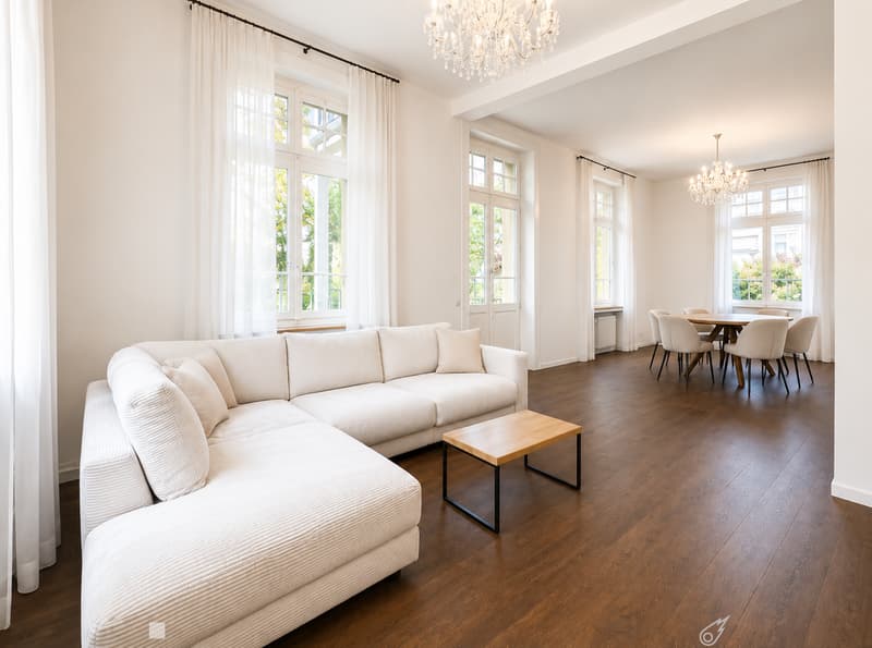 Bright living room with white walls, wooden flooring, large windows with white curtains, a white sectional sofa, a small wooden coffee table, a dining area with a table and chairs, and two chandeliers.