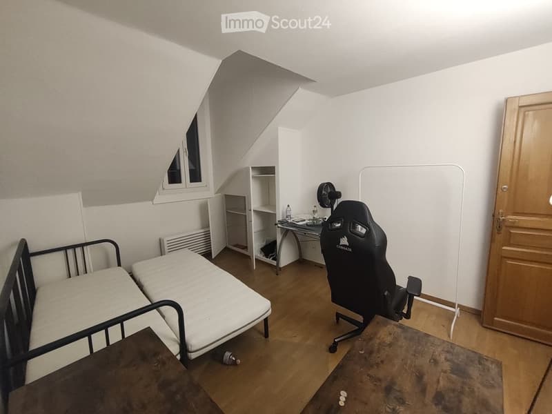A small, furnished room with a sofa bed, a gaming chair, a small table, a fan, and a shelf. The room has wooden flooring and white walls.