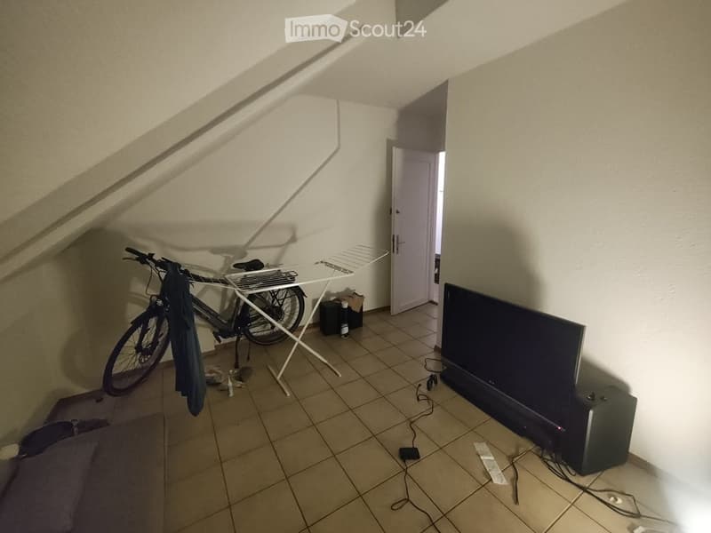 A small, furnished room with a tiled floor, a television on a stand, a bicycle leaning against the wall, and an ironing board. The room has a slanted ceiling and an open doorway leading to another space.