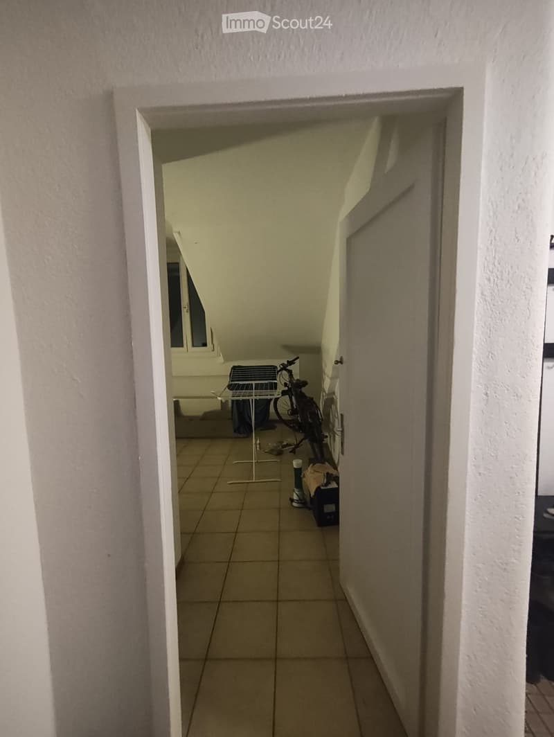 A narrow hallway with tiled flooring and white walls, leading to a room with a bicycle and a drying rack visible in the background.
