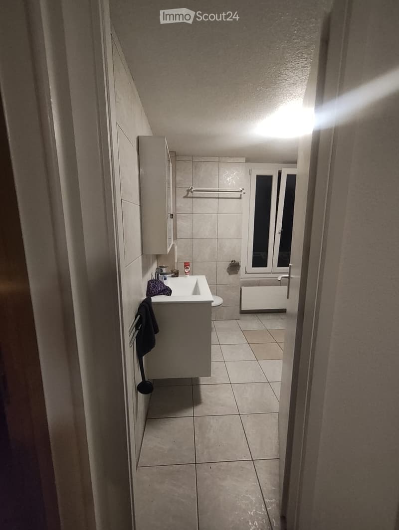 Modern bathroom with beige tiles, a white sink, a mirror, a towel hanging on the wall, and a toilet visible in the background.