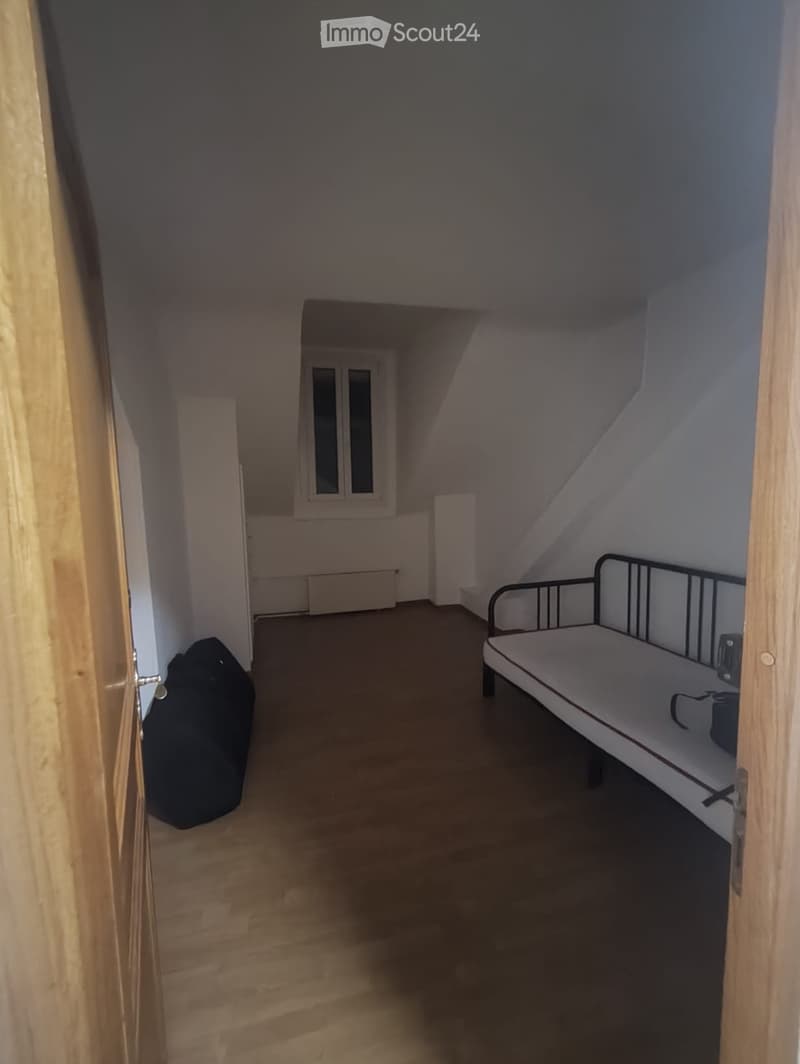 Small attic bedroom with wooden floor, slanted ceiling, single bed, radiator, backpack, and window with white frame.