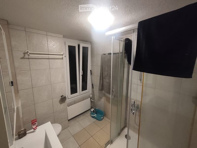 Modern bathroom with tiled walls and floor, featuring a toilet, sink, shower, towel, mop, and toothpaste on the sink.