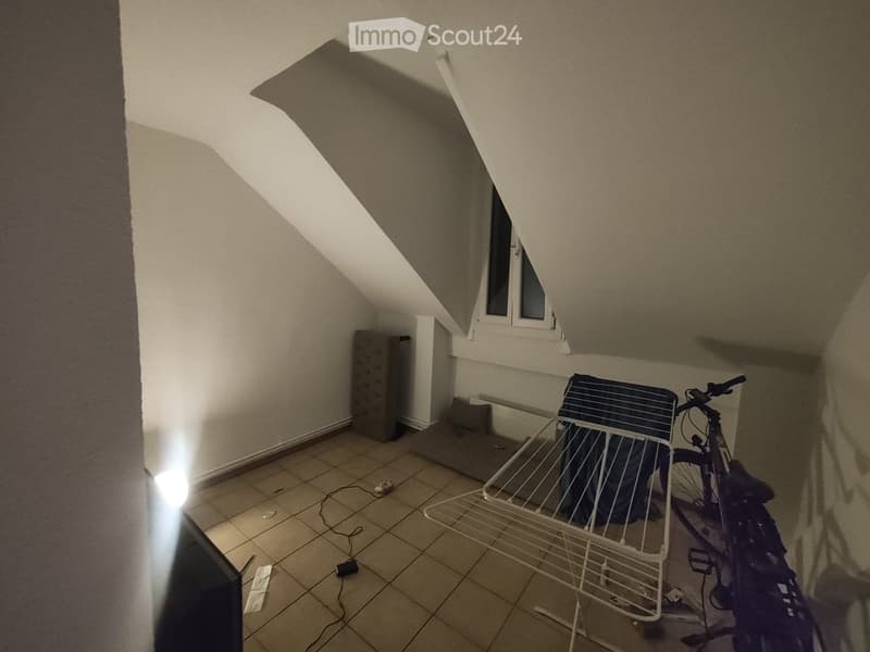 An attic room with slanted ceilings, tiled floor, and a window. Contains a bicycle and a drying rack.