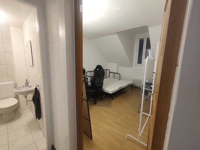 A small, furnished room with a bed, chair, and radiator, connected to a tiled bathroom with a sink and toilet.