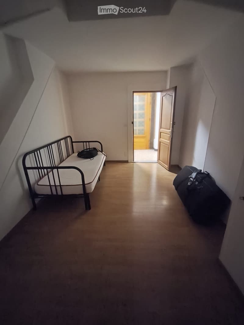 A small, furnished room with a single bed, a suitcase, and an open doorway leading to another room.