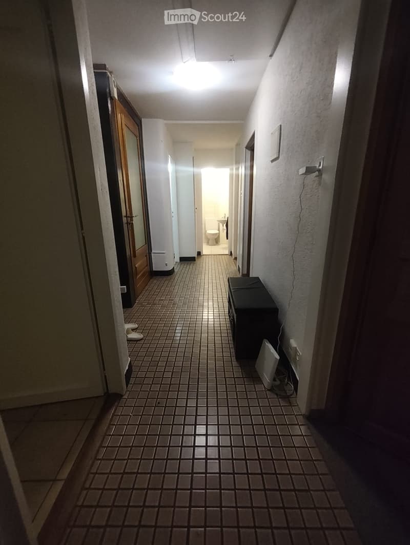 A narrow hallway with tiled flooring, white walls, and a visible bathroom at the end. The hallway features a wooden door on the left and a black cabinet on the right.