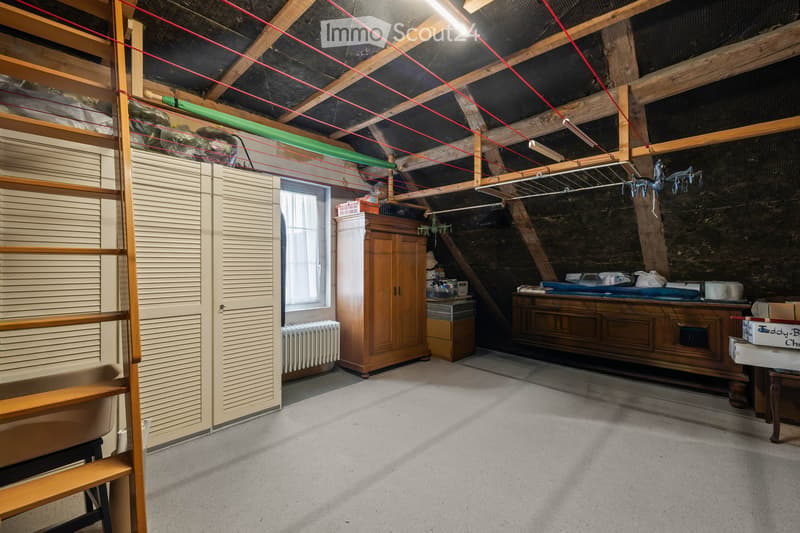 Attic space with wooden beams, exposed wiring, a window, a wooden cabinet, a radiator, a table, and various storage items.
