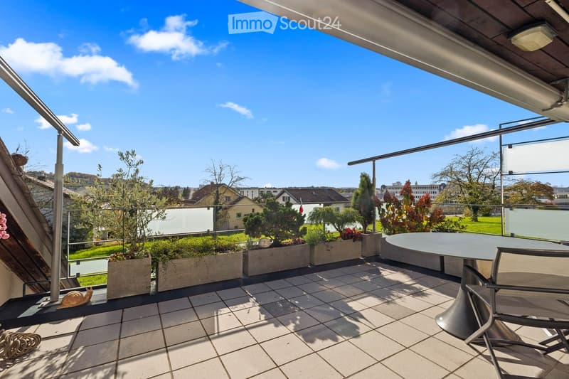 A modern balcony with tiled flooring, glass railing, and several potted plants arranged along the edge. A round table and a single chair are positioned near the railing, offering a view of the surrounding greenery and cityscape.