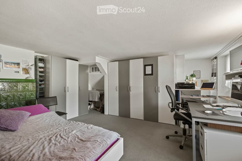 Modern bedroom with a bed, desk, chair, laptop, printer, and a wardrobe. The room features a grey carpet, white walls, and a green cabinet.