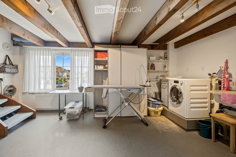 Well-lit laundry room with wooden beams, white washing machine, ironing board, table, shelves, and various laundry items.