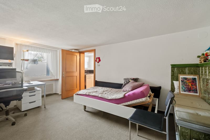 A well-lit bedroom with a single bed, desk, chair, and wardrobe. The bed has a pink blanket and pillows, and there is a framed picture on the wardrobe.