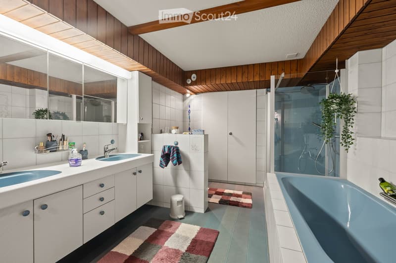 Modern bathroom with a double sink, large mirror, bathtub, and walk-in shower. Features wooden paneling on the ceiling, white tiles, and colorful bath mats.