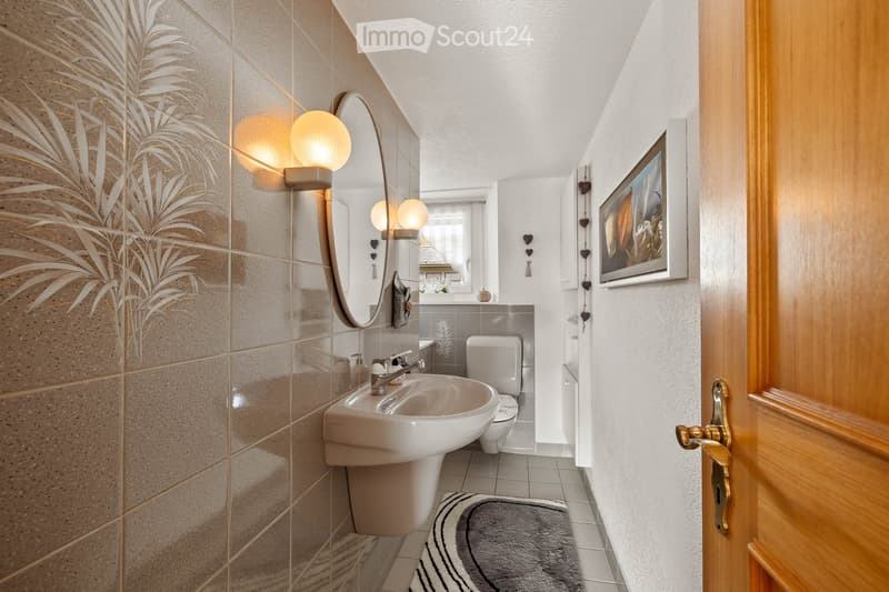 Modern bathroom with a white sink, chrome faucet, toilet, large mirror with integrated lighting, and a decorative picture frame on the wall. The floor is tiled, and a grey bathroom rug is placed in front of the sink.
