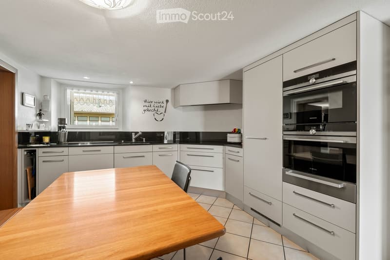 Modern kitchen with white cabinets, stainless steel appliances, a wooden dining table, and a window overlooking the street.