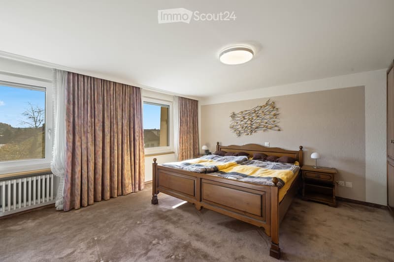 Cozy bedroom with a wooden bed, nightstand, and wall-mounted decorative art. Large windows with curtains provide natural light.