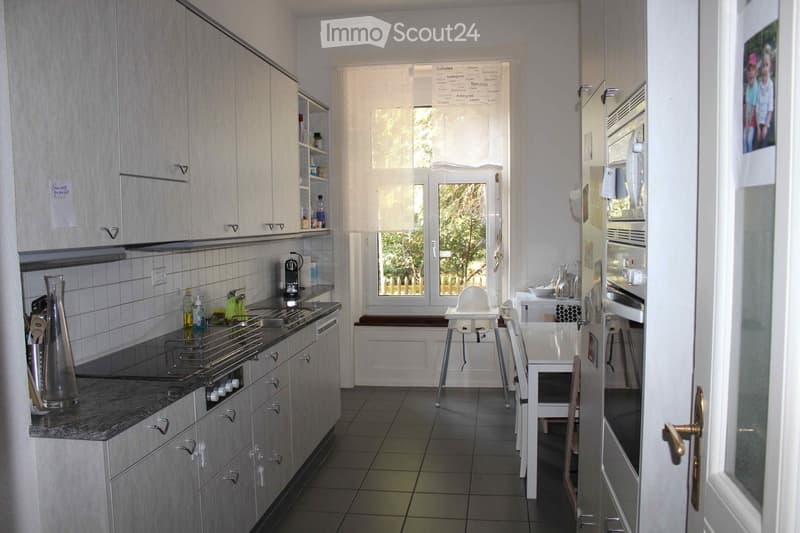 Modern kitchen with light grey cabinets, granite countertop, built-in stove, dishwasher, and refrigerator. Includes a small dining area with a table, chairs, and a baby chair. A window with blinds offers natural light and a view of greenery outside.