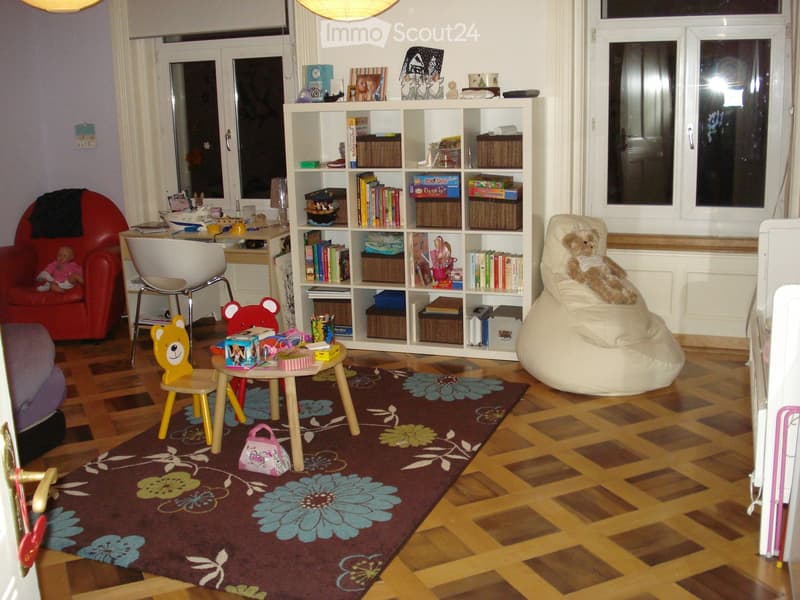 A well-lit children's room with wooden flooring, a large bookshelf filled with books and toys, a red armchair, a white beanie chair, a small table with toys, and a colorful rug with floral patterns.