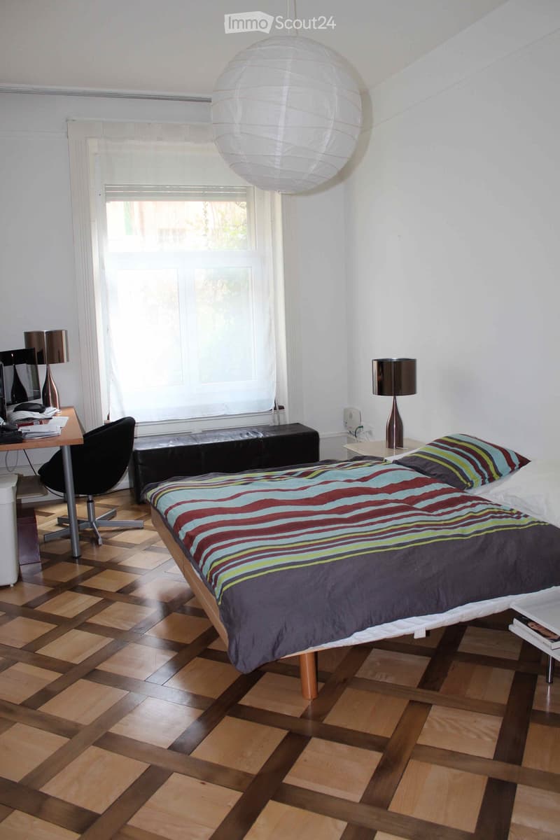 A modern bedroom with a wooden floor, a large bed with a colorful striped duvet, a desk with a chair, and a lamp on a side table. There is a large window with blinds and a hanging paper lantern light fixture.