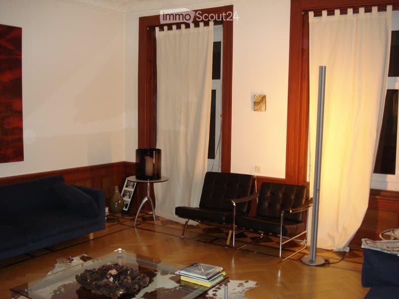 Living room with wooden flooring, a blue sofa, two black leather chairs, a glass coffee table with books and a decorative item, a small round table with a vase and candle, and a tall floor lamp.