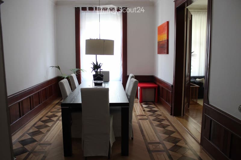 Dining room with a long wooden table, six chairs, a pendant lamp, a red stool, and a painting on the wall. The room has patterned wooden flooring and large windows with curtains.