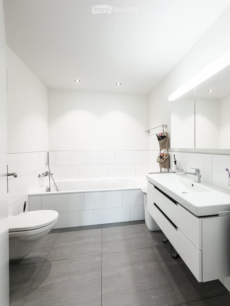 Modern bathroom with white tiles, a bathtub, toilet, sink, and a large mirror. Features include built-in cabinets, a shower head, and a sleek faucet.