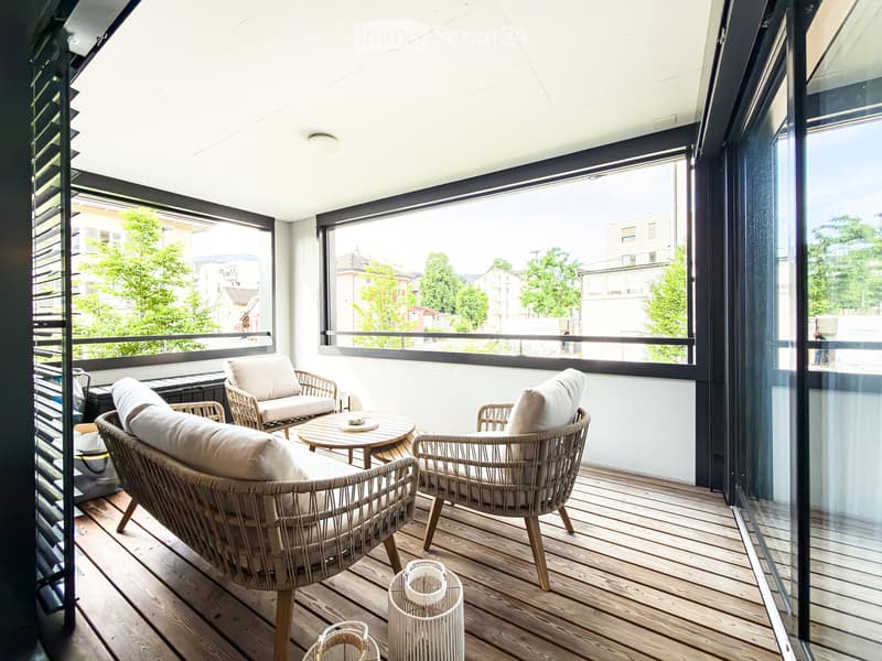 A modern balcony with wooden flooring, large windows, and wicker furniture including a sofa, chairs, and a small table.