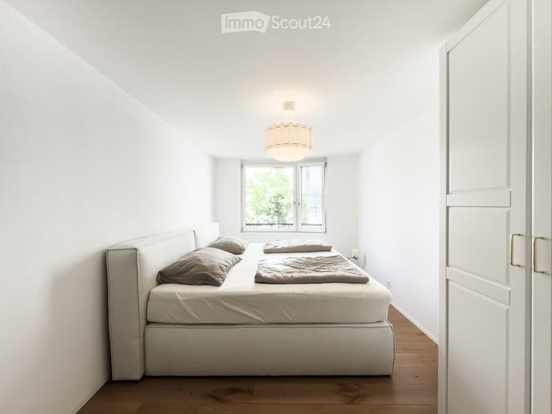Minimalist bedroom with a single bed, pillows, and a blanket, featuring a pendant light and a window with a view of greenery.