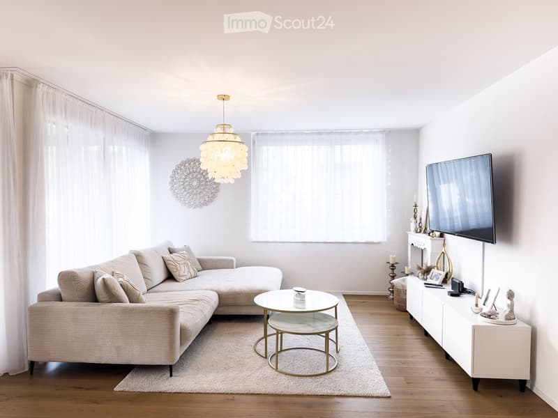 Modern living room with a beige sectional sofa, a round coffee table, a TV on a white cabinet, and decorative items on the cabinet. The room features wooden flooring, large windows with white curtains, and a hanging chandelier.
