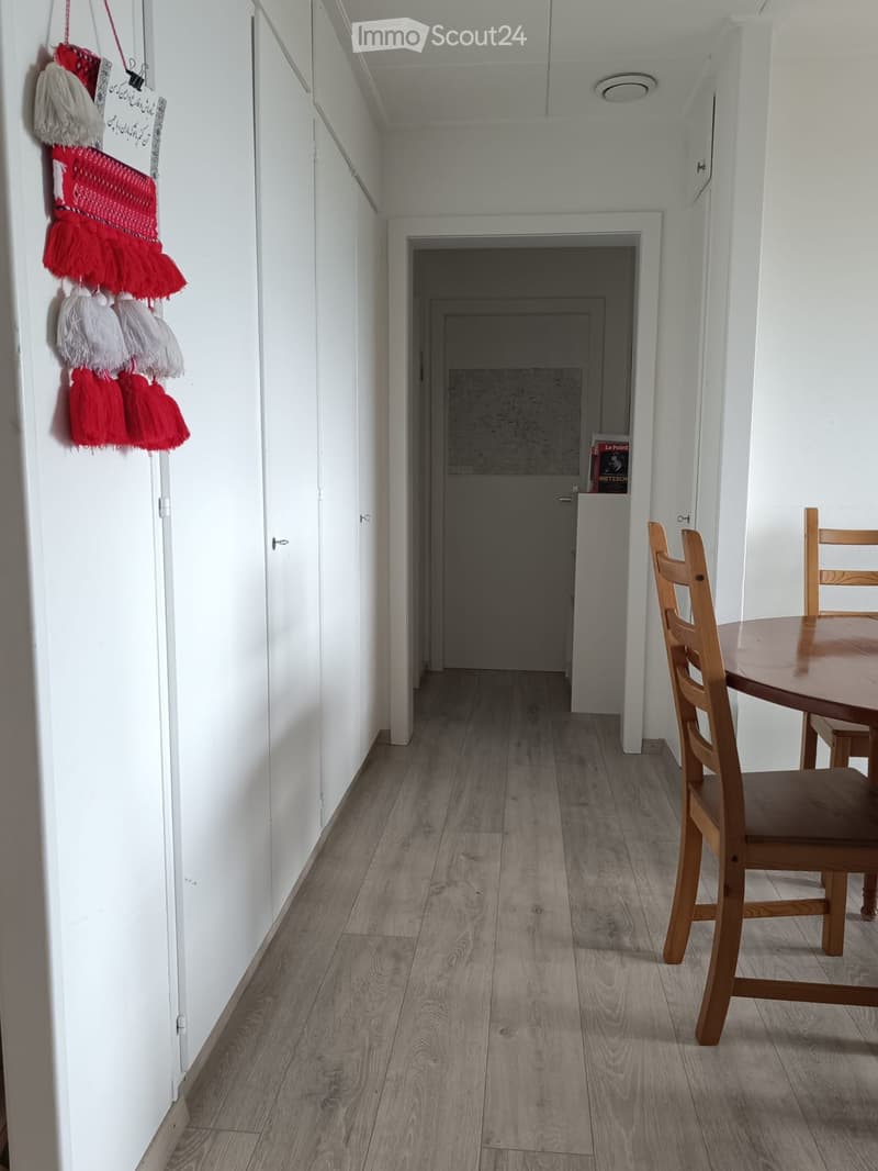 A narrow hallway with light wooden flooring and white walls, featuring a small table and chair in the corner and a decorative hanging ornament on the wall.