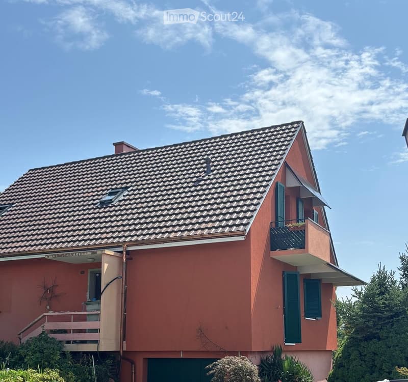 Two-story house with red exterior, grey tiled roof, balcony on the upper level, and green shutters.