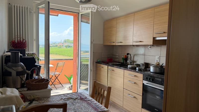Modern kitchen with light wooden cabinets, stainless steel appliances, and a dining area. Large sliding doors open to a balcony with a scenic view of a green field and mountains.