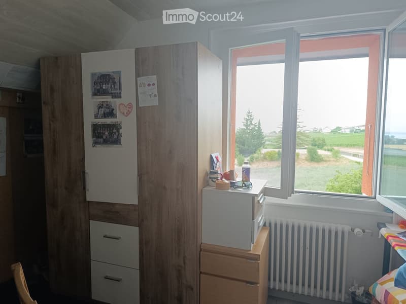 A small, furnished room with a refrigerator, wooden cupboard, small table with items, and a window offering a view of the countryside.