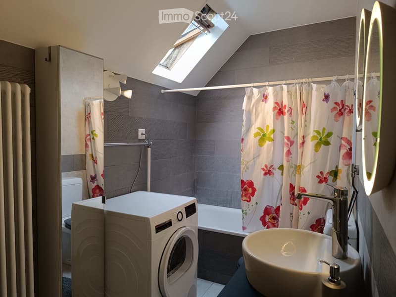 Modern bathroom with a skylight, grey tiled walls, a white sink, a bathtub with a floral shower curtain, and a washing machine.
