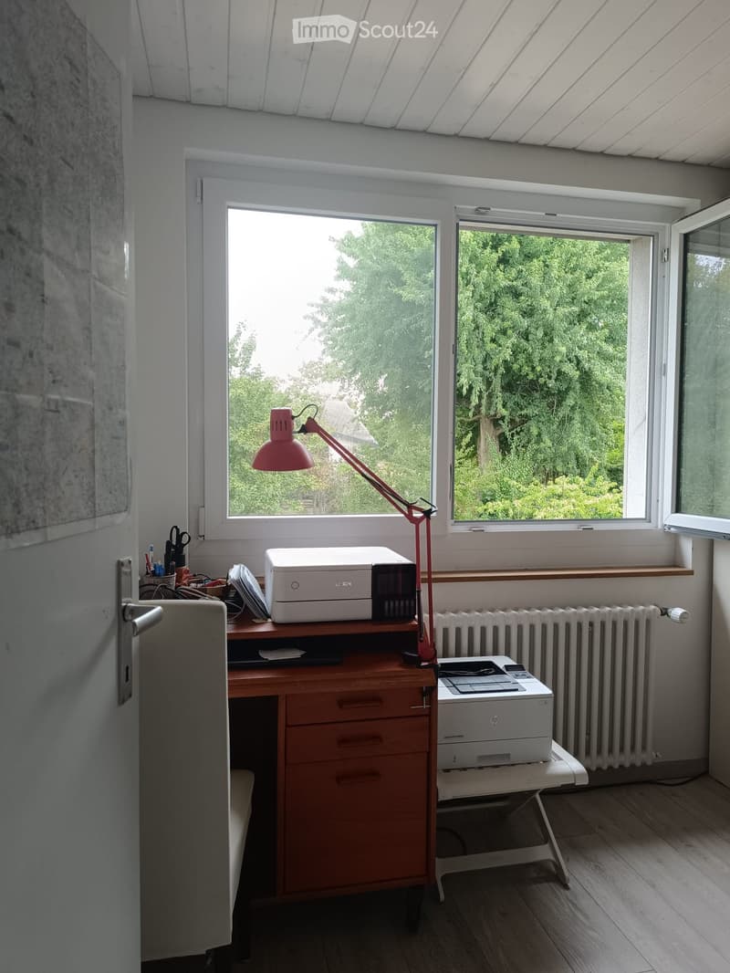Bright room with wooden flooring, large windows, a desk with a printer, a red lamp, and a radiator.