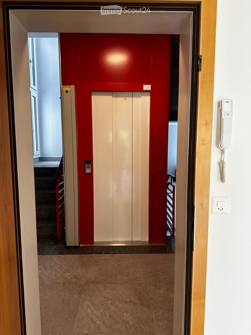 Hallway with elevator doors, red accent wall, tiled floor, intercom system, and a visible staircase area.