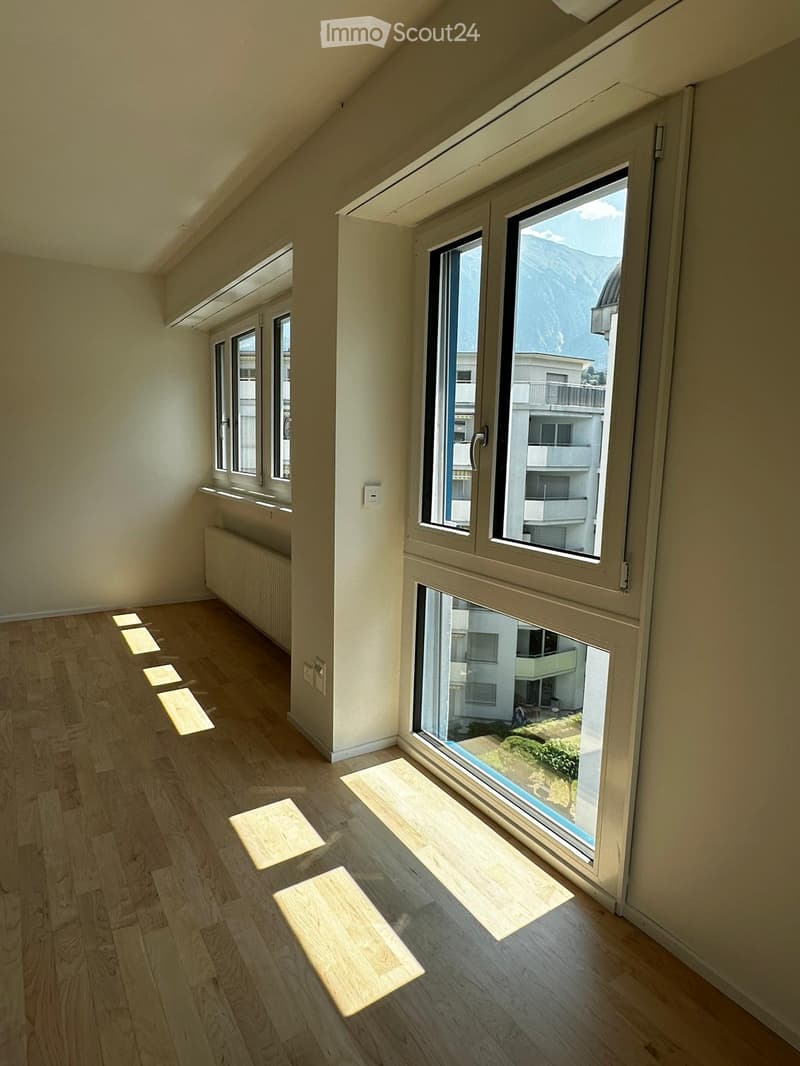 Empty room with wooden flooring, large windows, and natural light. The room has a slanted ceiling and a view of mountains and neighboring buildings.