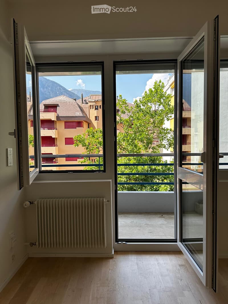Empty room with wooden flooring, large windows offering a scenic mountain view, radiator, and open sliding doors leading to a balcony.