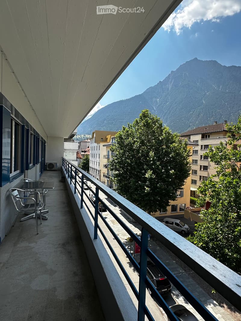 Balcony with a view of mountains and city buildings, equipped with a table and chairs.