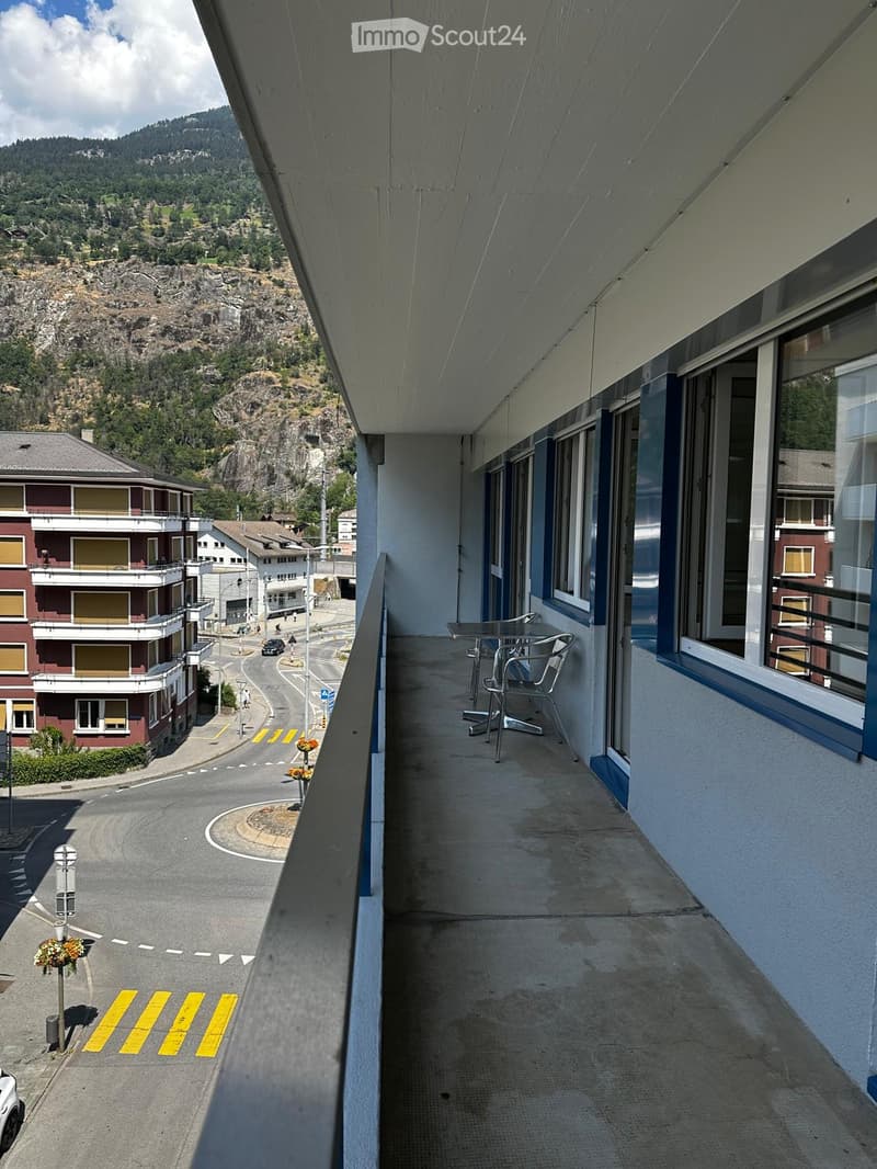 Balcony with a table and chairs, overlooking a street and surrounding mountains.