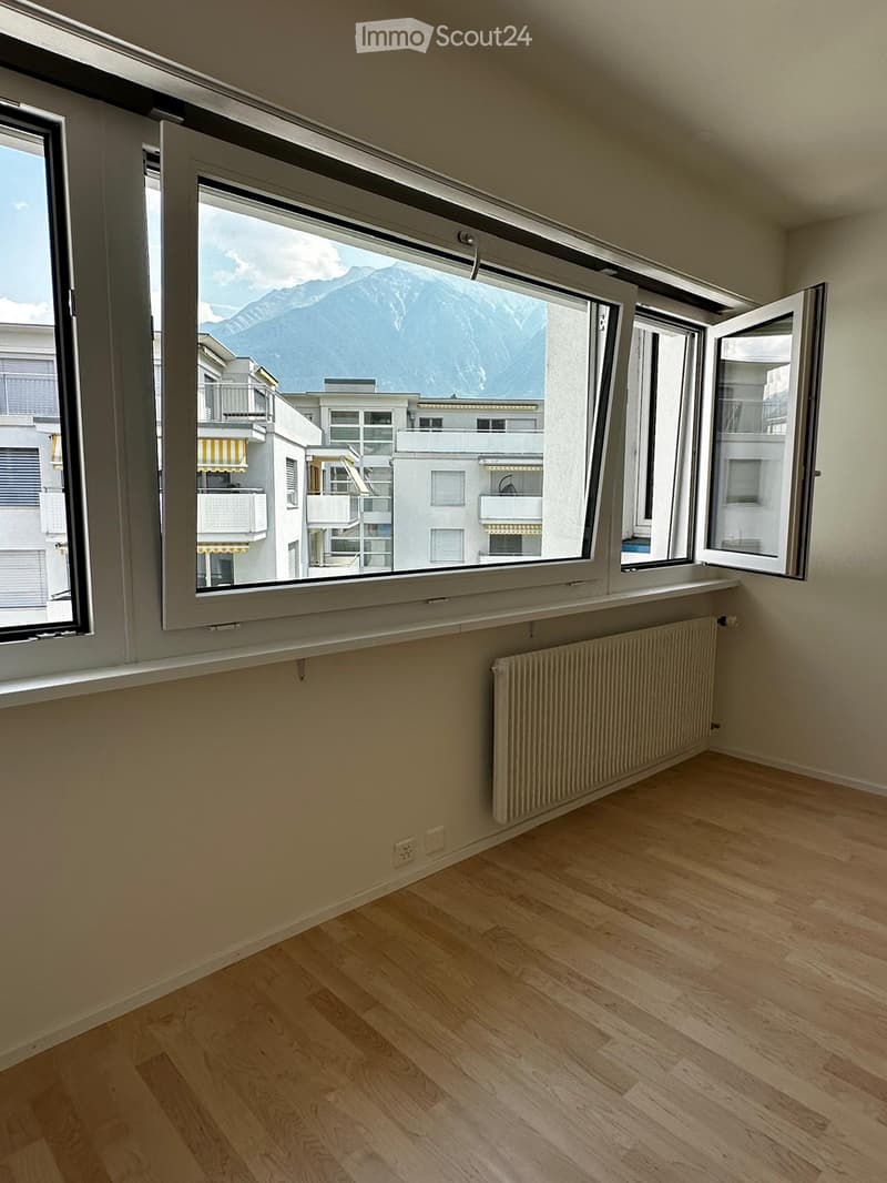 Empty room with wooden flooring, large windows offering a mountain view, radiator, and electrical outlets.