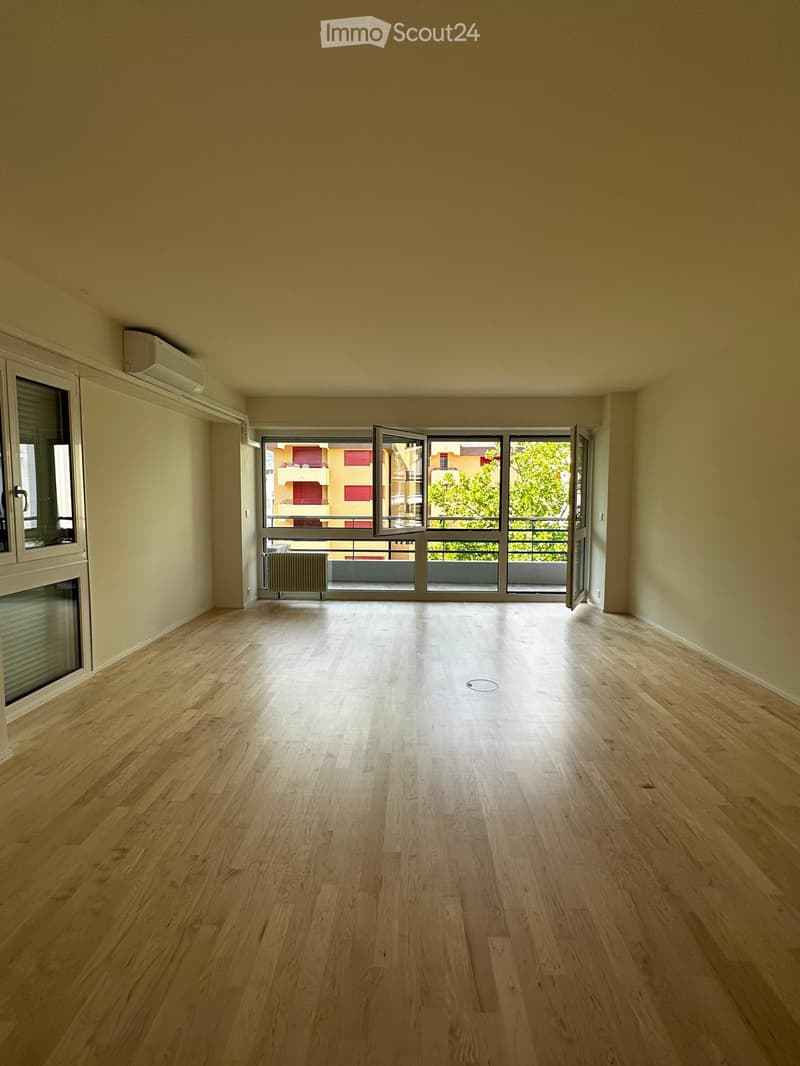 Empty room with light wooden flooring, large windows opening to a balcony, and neutral-colored walls.
