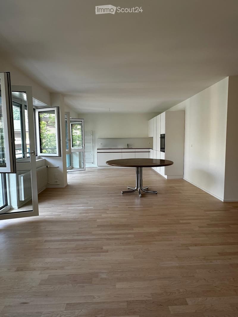 Open-plan space with light wooden flooring, white walls, and a single round table near the kitchen area. Large windows and sliding doors provide natural light and access to the outdoors.