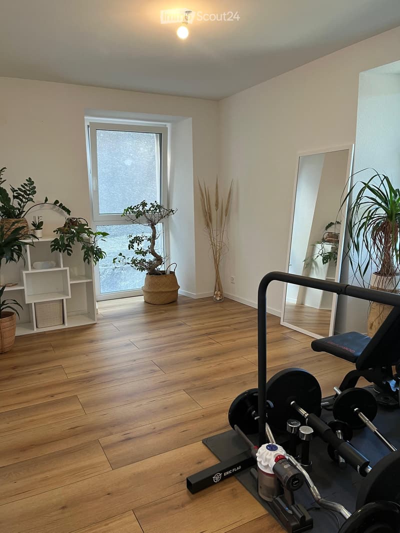 Well-lit room with wooden flooring, large window, mirror, shelf with potted plants, and fitness equipment including a weight machine and weight plates.
