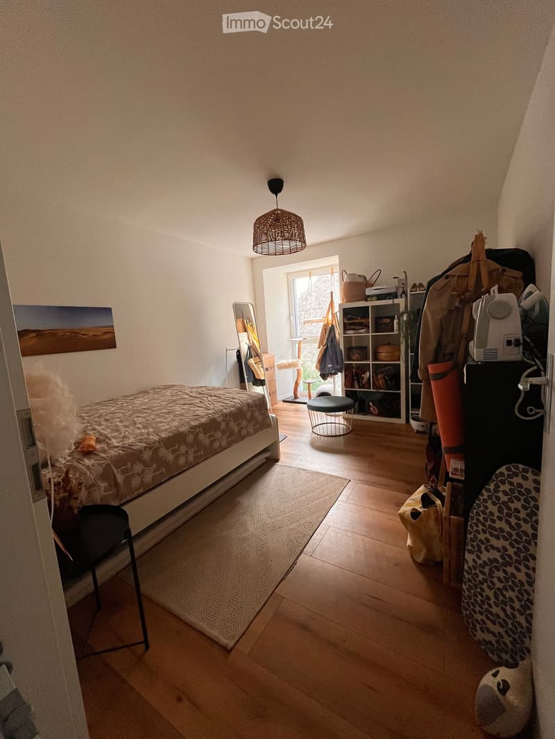 Cozy bedroom with wooden flooring, a bed with a patterned duvet, a chair, a shelf with various items, a small round stool, a mirror, and a fan.