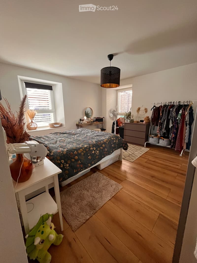 Cozy bedroom with wooden flooring, a double bed with a patterned duvet, a nightstand with a lamp, a mirror, a fan, a dresser, a clothing rack, and decorative items including a vase and a rug.