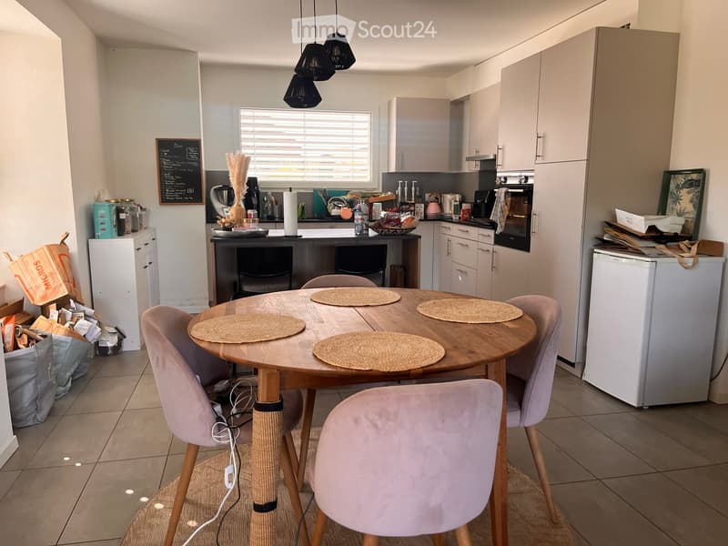 Modern kitchen with a round wooden dining table, four pink chairs, and various appliances including a refrigerator, oven, and coffee maker. The kitchen features white cabinets, a black countertop, and a window with blinds.
