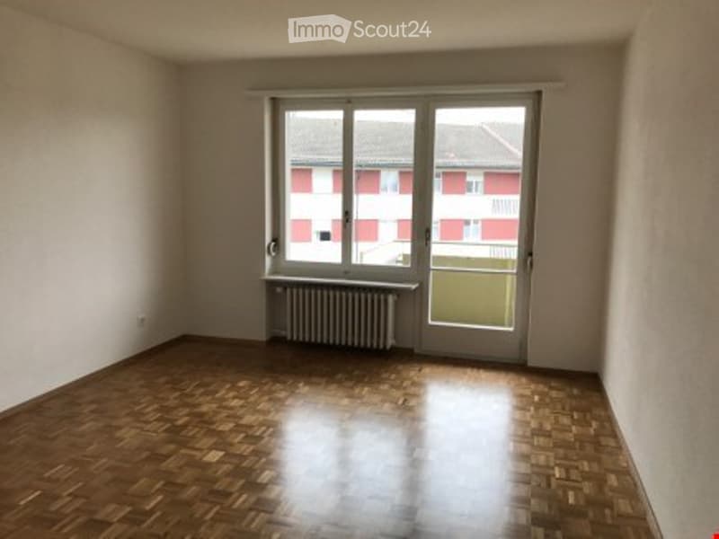 Empty room with wooden flooring, large window, radiator, and door leading outside.