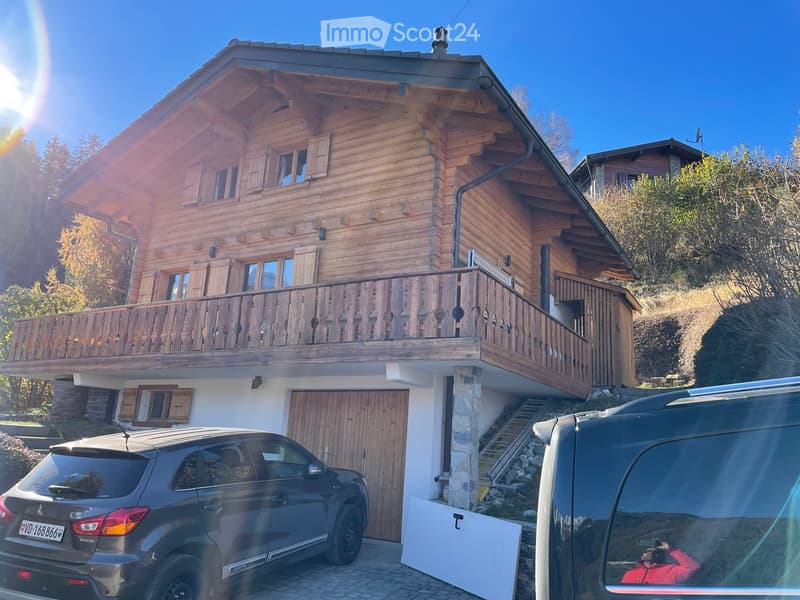 A two-story wooden house with a balcony, garage, and two parked cars in front.